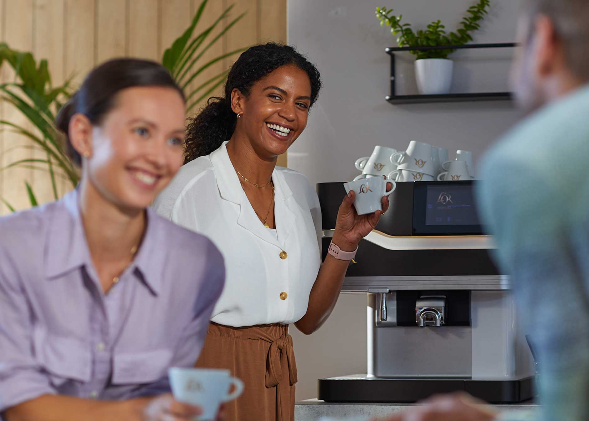 Tre personer diskuterar vid en kaffemaskin, var och en med en kopp i handen.