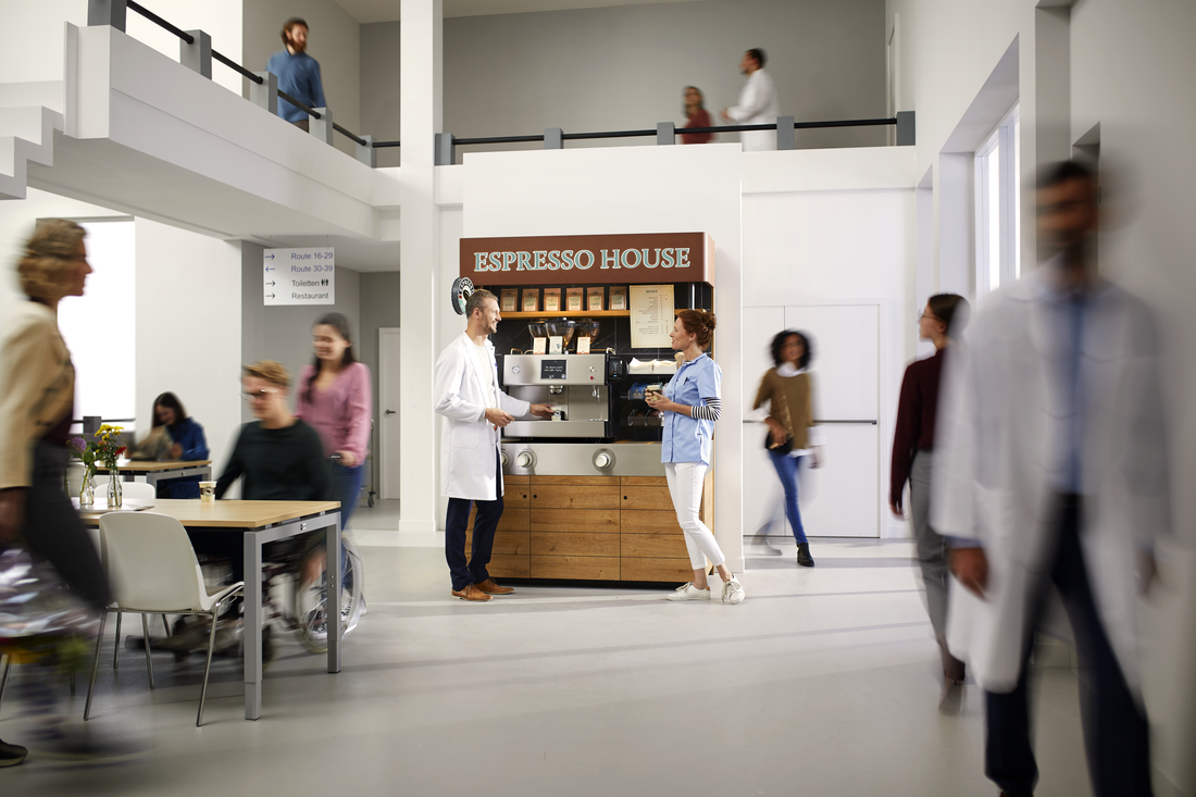 Kaffestation med skylten 'ESPRESSO HOUSE' i en ljus vårdmiljö där en man i vit rock samtalar med en kvinna med kaffekopp, omgiven av människor inklusive en person i rullstol.