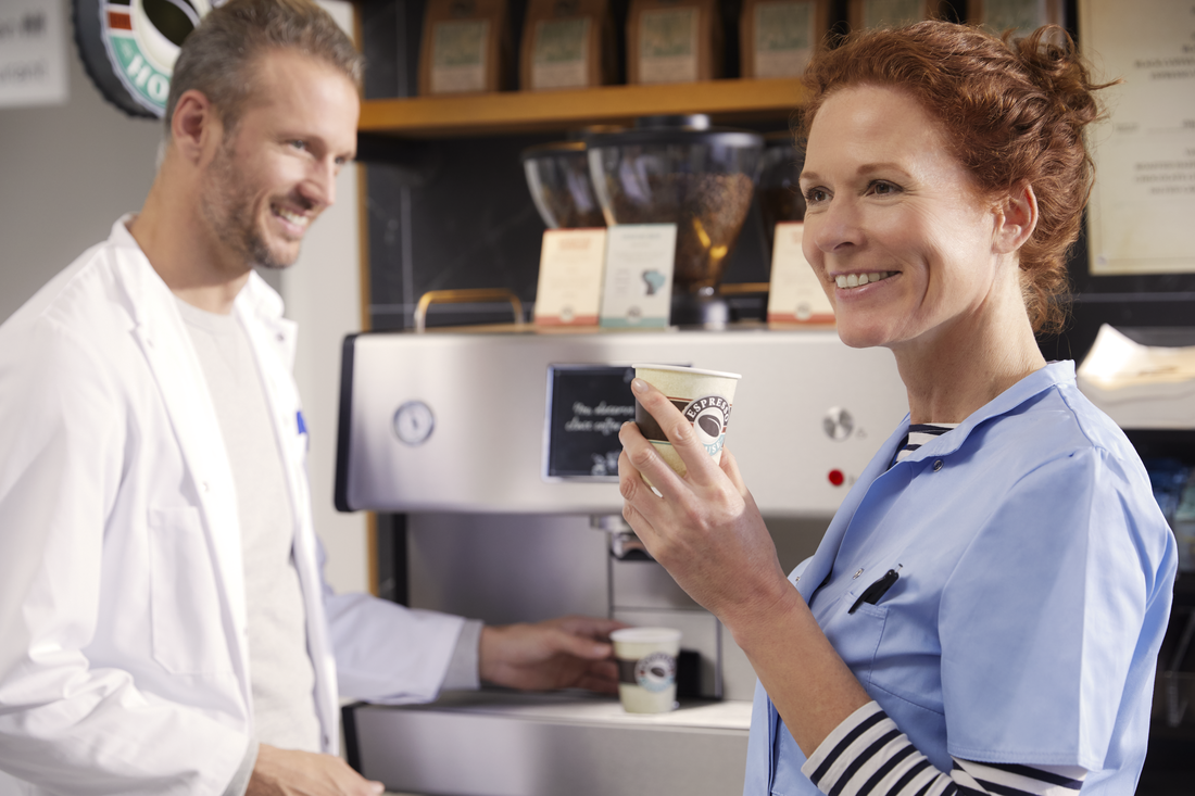 Två vårdpersonal, en i vit rock och en i blå scrubs, står vid en kaffemaskin och håller pappersmuggar i en pausmiljö inom vården.